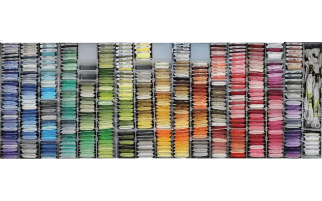 Des fils à broder DMC sont enroulés sur cartonnette et rangés dans des boîtes transparentes. Ils sont classés par couleur, du bleu-violet à gauche aux couleurs neutres à droite.