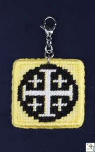 Réalisation sur demande. Un bijou fantaisie représentant une croix de Jérusalem blanche sur un fond noir rond entouré de jaune or est photographié sur un fond bleu marine. Les côtés gauche de la croix sont soulignés de jaune or.