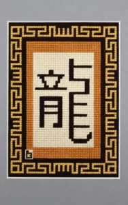 Broderie contemporaine. Entourée d'un passe-partout gris à biseau blanc, la tapisserie représente l'idéogramme chinois dragon en couleur ébène sur un fond rectangulaire écru. Ce dernier est entouré par un rectangle de couleur chêne, lui même entouré par une frise couleur pin sur fond ébène représentant une farandole de dragons. La signature est en bas à gauche.